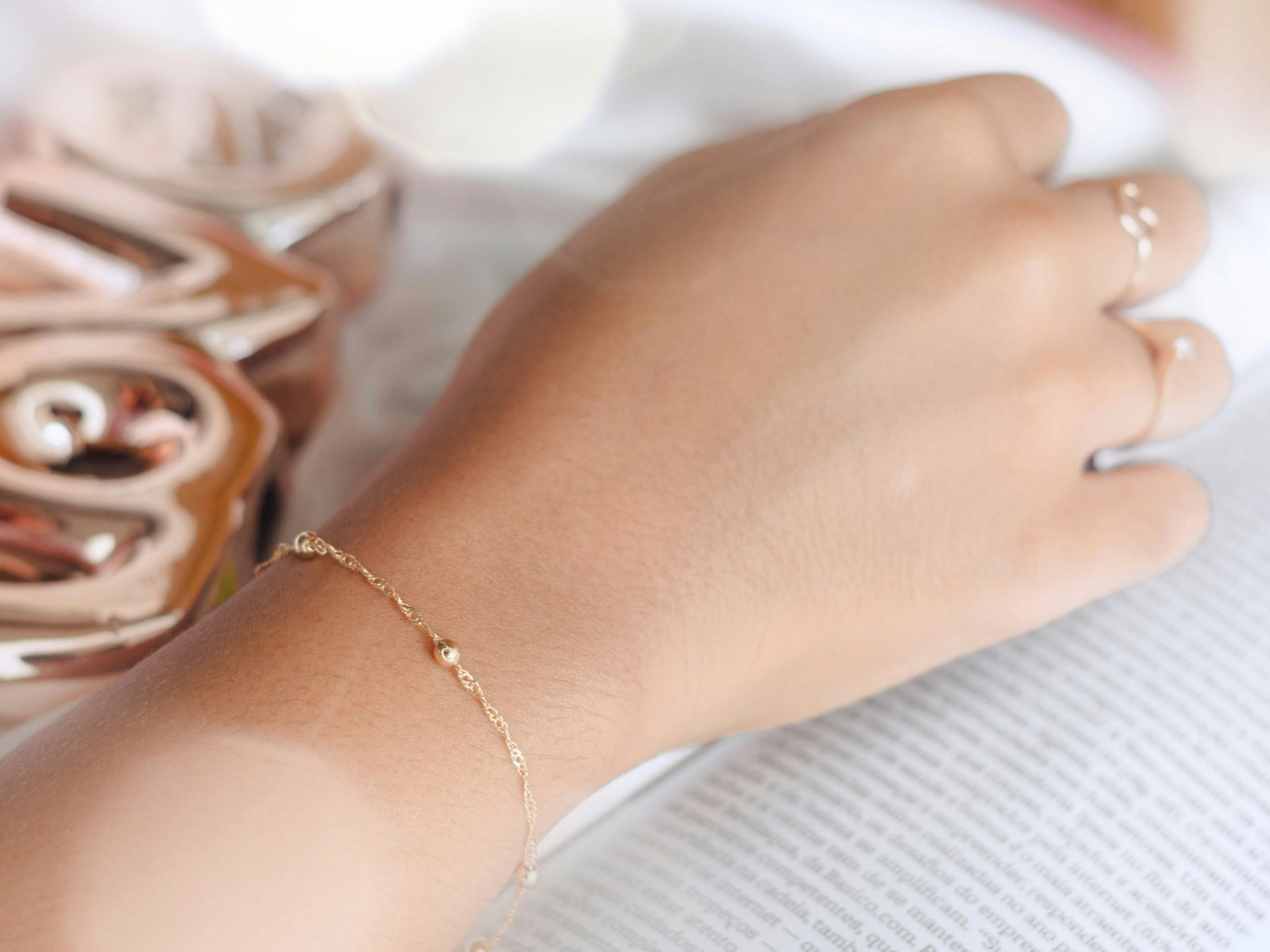 A delicate gold permanent bracelet with a small charm on a wrist, resting beside an open book.