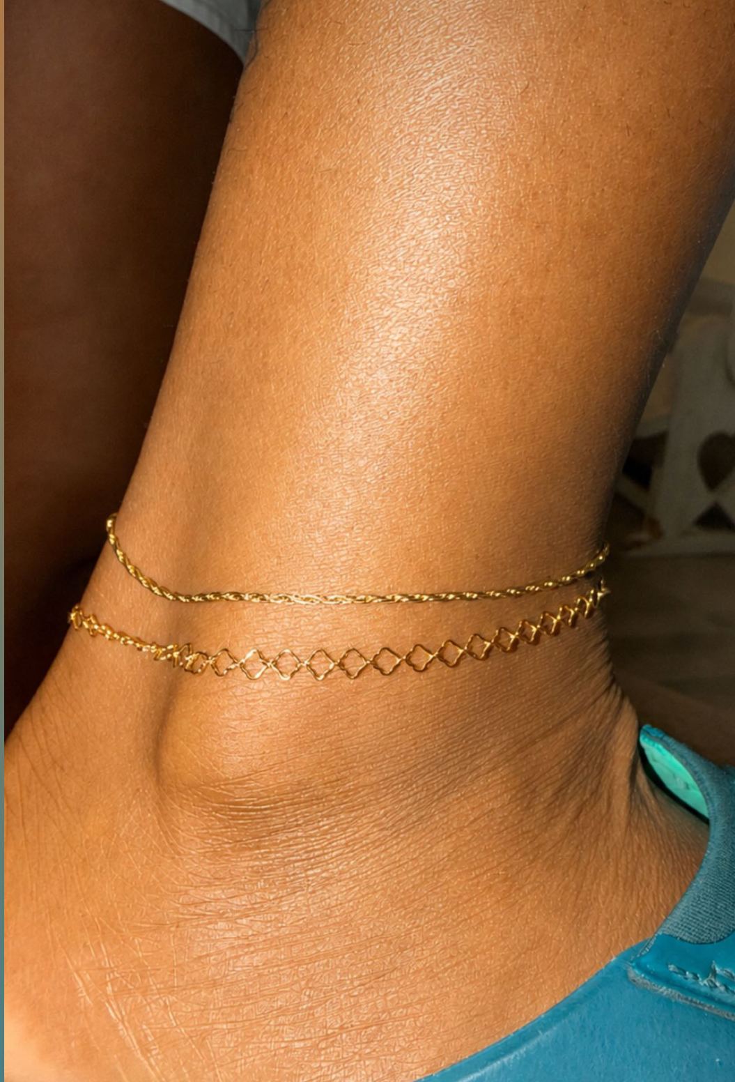 Two gold permanent anklets, a twisted rope chain and an open-link chain, layered on an ankle.