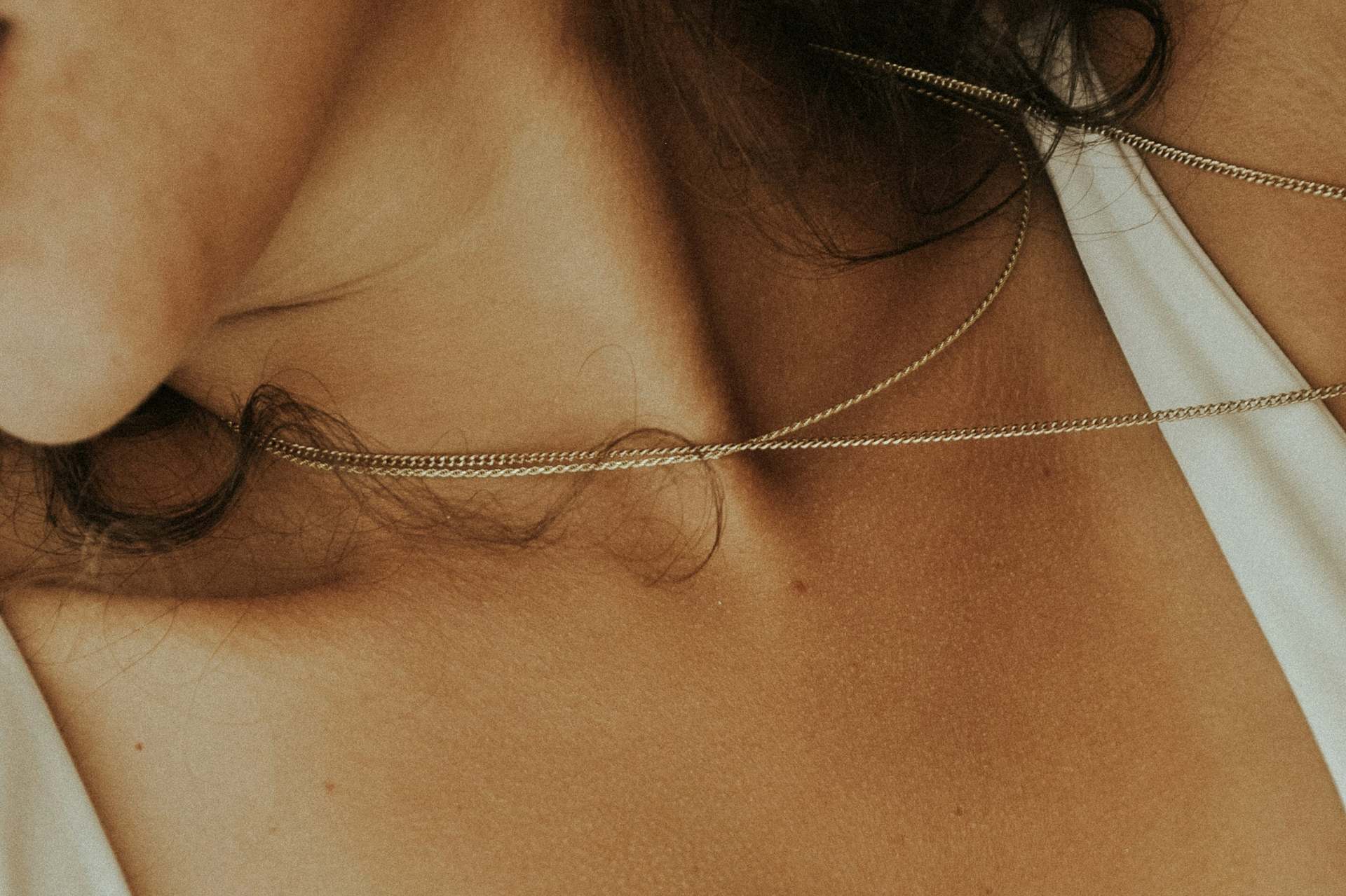 A fine gold curb-chain permanent necklace welded at a woman's collarbone, her dark hair falling loosely around it.