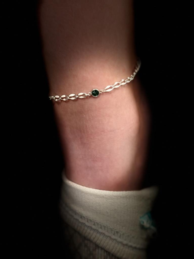 A silver permanent anklet with flat oval links and a round dark green stone connector, worn above a cream sock.