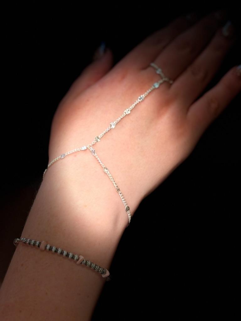 A silver permanent hand chain running from a wrist bracelet across the back of the hand to a ring on the middle finger.