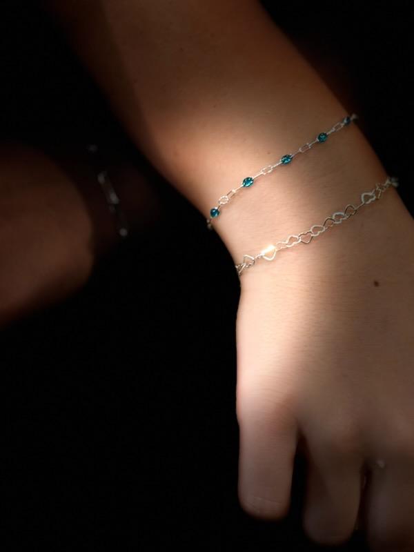 Two silver permanent bracelets on a wrist, one with teal enamel beads and one made of small open heart links.