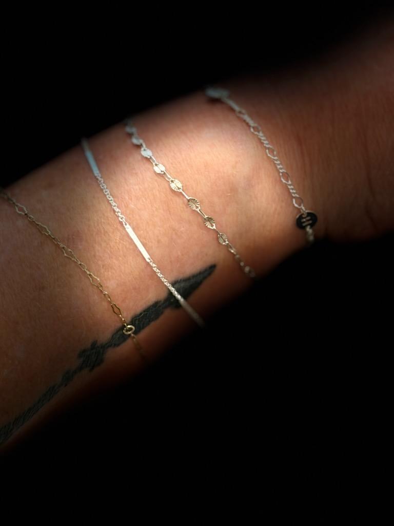 Four fine permanent bracelets in different chain styles stacked along a forearm, lit against a dark background.